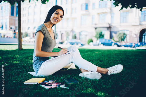 Smiling young woman in casual wear working on project for college spending time in capus,attractive cheerful girl making drawing in sketchbook sitting on lawn during free time in park on summer day photo