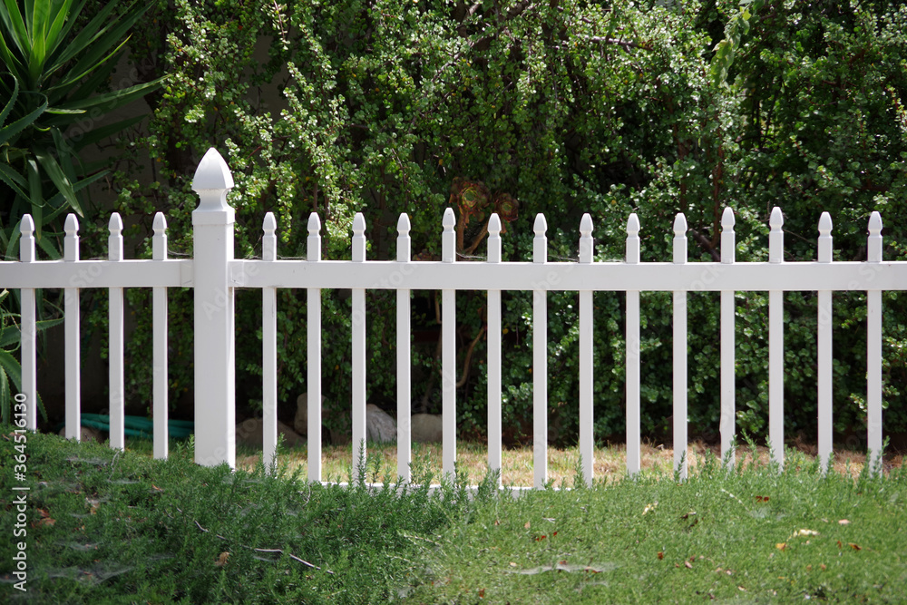 Close view of a section of a white picket fence with green bushes behind Close view of a section of a white picket fence with green bushes behind