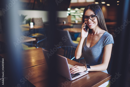 Happy woman calling to customer service for consultancy about application on laptop computer, successful hipster girl in spectacles enjoying time in coworking space while communicated via cellphone