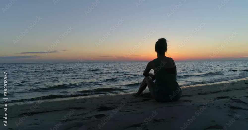 Female model sitting on a beach watching a beautiful sunset over the water, aerial drone shot