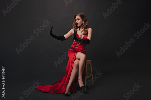 Lady in red long dress sitting on black background. Superstar in luxury outfit posing on isolated backdrop