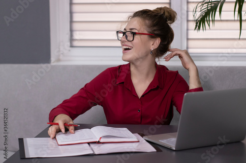 Happy, beautiful blonde girl in a red shirt and red glasses works in the office or at home. remote work or study. With a gray laptop. In a gray room on a gray sofa at a gray table. High quality photo