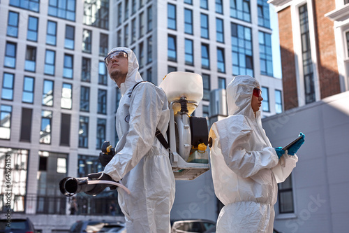 young confident specialists in hazmat protective suits cleaning and disinfecting coronavirus cells epidemic, pandemic health risk. in public places of city