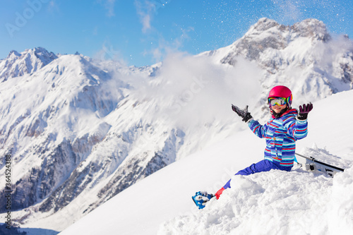Happy girl sit in snow on top of the mountain and lift hand in ski outfit, color mask, pink helmet