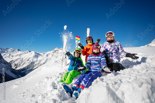 Large group of ski children in colorful outfit throw snow in the air sitting together over mountain tops view