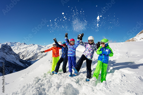 Fun portrait of a group of ski kids in colorful outfit throw snow in the air sitting together over mountain tops view