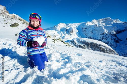 Ski little girl hold snow in heart shape smiling with mountain summit on background standing on her knees