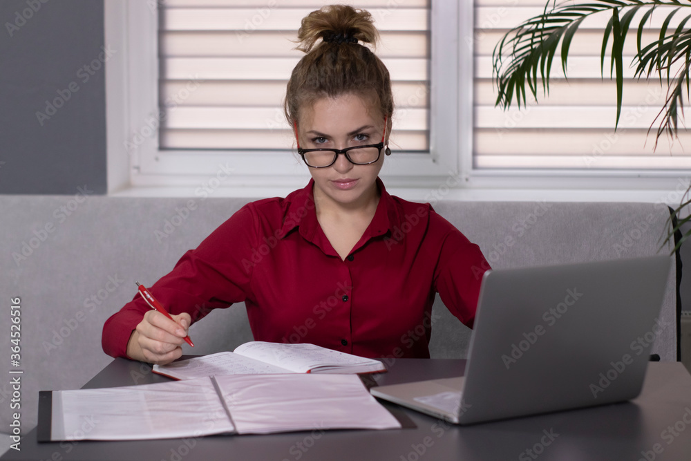 Pensive, beautiful blonde girl in a red shirt and red glasses works in the office or at home. remote work or study. With a gray laptop. In a gray room on a gray sofa at a gray table Pensive, beautiful blonde girl in a red shirt and red glasses works in the office or at home. remote work or study. With a gray laptop. In a gray room on a gray sofa at a gray table