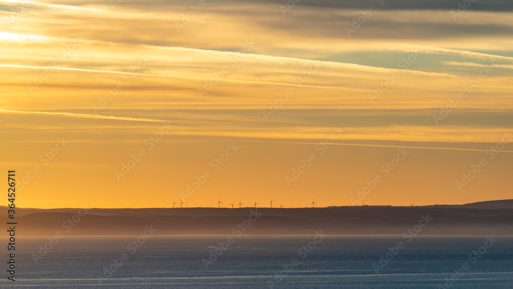 Wind farm during very golden hour with wind turbines at the cliff top at the horizon above calm sea under golden morning sky