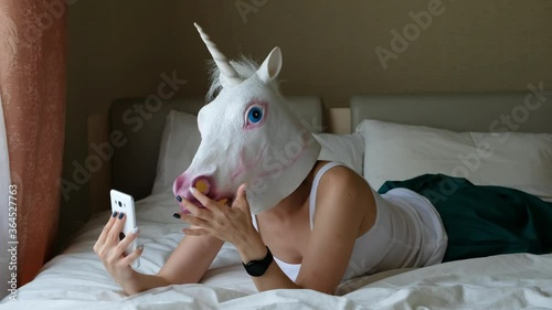 Woman with a head mask of unicorn talking to web camera on smart phone on a bedroom. Social media concept. Weird funny video