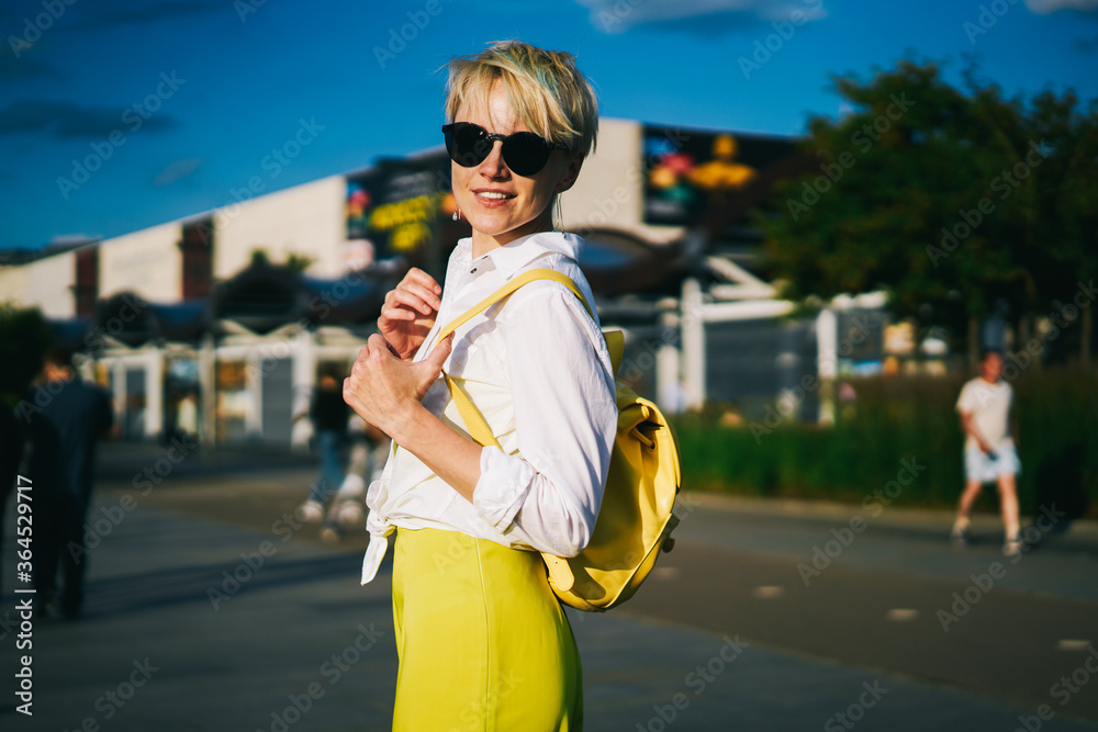 Fototapeta premium Portrait of cheerful fashionable young woman with trendy yellow backpack and stylish black sunglasses standing outdoors in city centre while looking at camera during walk in good sunny weather