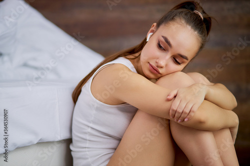 sad melancholic woman is listening to depressive music in headphones, sits on floor near bed, looking down