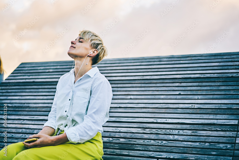 Young woman enjoying favourite music in earphones connected to telephone sitting on wooden bench in recreation time outdoors in urban setting.Copy space area for your advertising text message - obrazy, fototapety, plakaty 