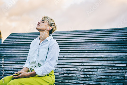 Young woman enjoying favourite music in earphones connected to telephone sitting on wooden bench in recreation time outdoors in urban setting.Copy space area for your advertising text message