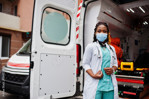 African american female paramedic in face protective medical mask standing in front of ambulance car. photo