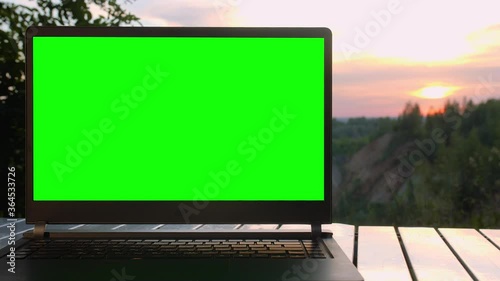 Black laptop with a green screen is on a folding table at sunset at top of mountain.