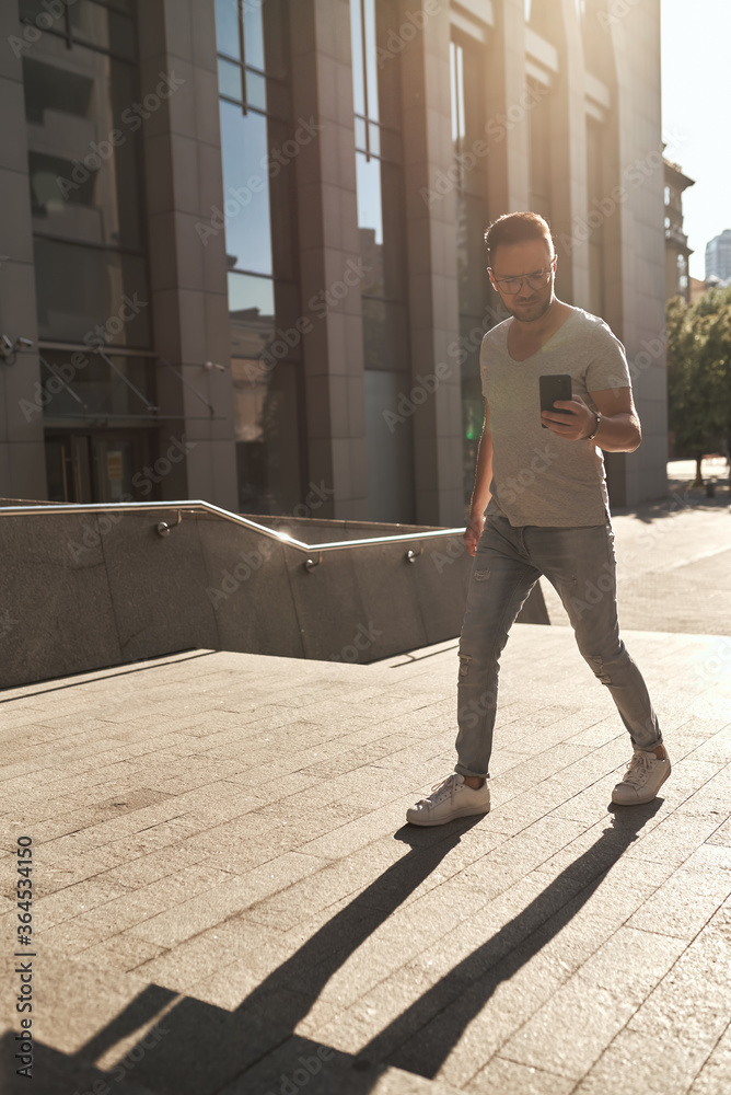 stylish guy, wearing glasses, climbs the steps to the office check-in with a phone in his hands. light jeans and fashionable gray T-shirt. beautiful contour sunlight stylish guy, wearing glasses, climbs the steps to the office check-in with a phone in his hands. light jeans and fashionable gray T-shirt. beautiful contour sunlight