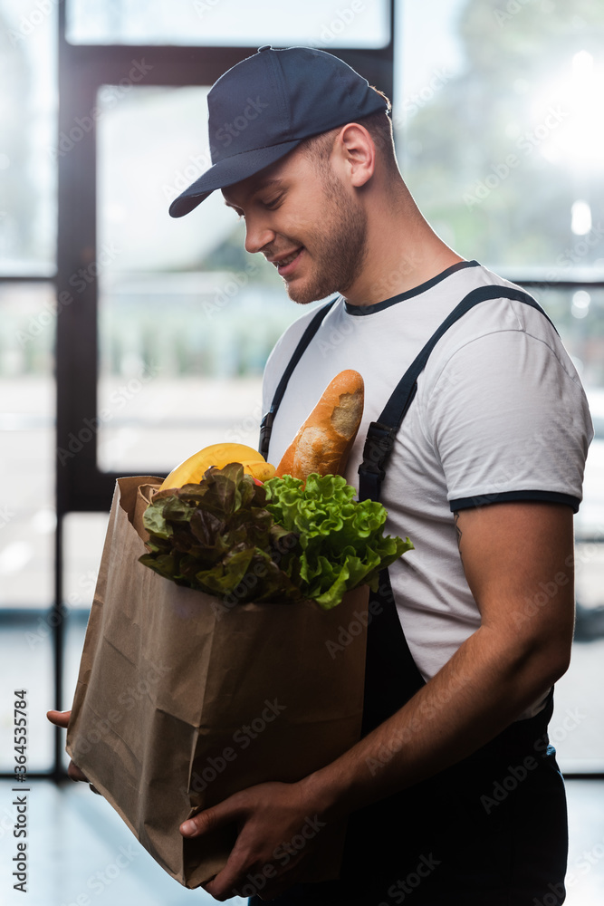 cheerful delivery man in cap looking at paper bag with groceries cheerful delivery man in cap looking at paper bag with groceries