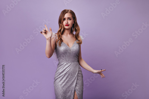 Lady with wavy hair pathetically looks into camera. Curly blonde woman in silver festive dress posing on isolated purple backdrop photo