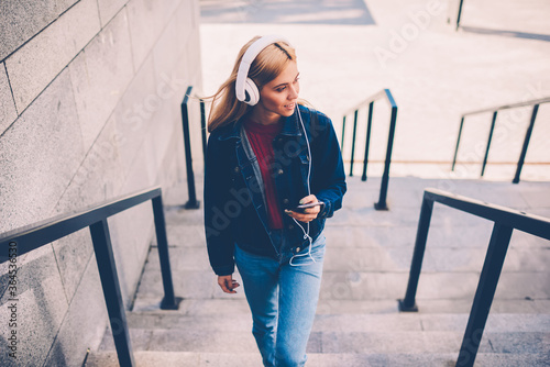 Stylish young woman in modern white headphones listening electronic music downloaded on own playlist walking in urban setting climbing stairs.Smart student learning foreign languages via audio lesson photo