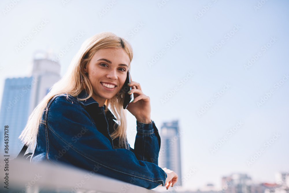 Portrait of carefree hipster girl with blonde hair smiling at camera while having mobile conversation with operator on smartphone device.Positive student talking in roaming on telephone enjoying trip