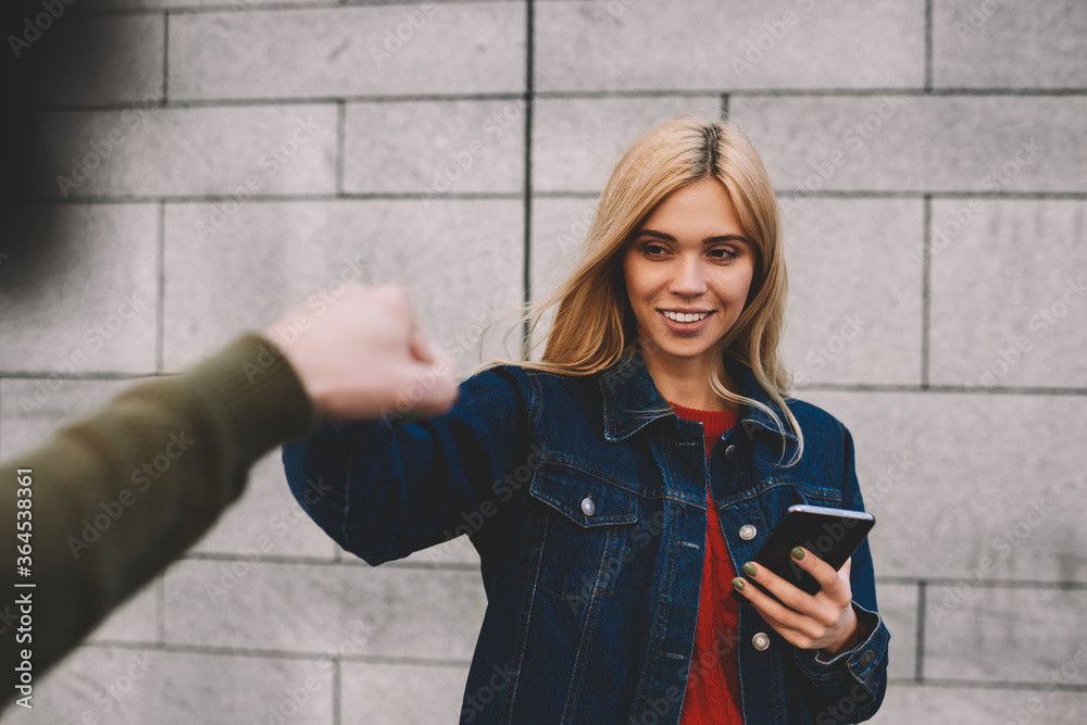Young charming smiling blonde dressed in stylish jeans clothes downloading application from network using cellphone and internet connection during giving knock fists to male best friend outdoors Young charming smiling blonde dressed in stylish jeans clothes downloading application from network using cellphone and internet connection during giving knock fists to male best friend outdoors