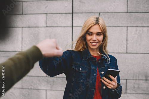 Young charming smiling blonde dressed in stylish jeans clothes downloading application from network using cellphone and internet connection during giving knock fists to male best friend outdoors