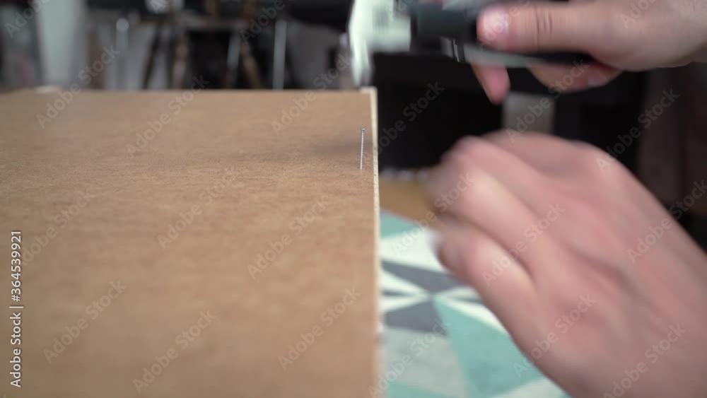 Handyman hammering a nail a on the board of a furniture to assemble it inside the living room of the house during day Handyman hammering a nail a on the board of a furniture to assemble it inside the living room of the house during day