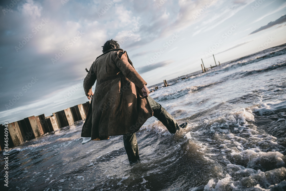 Naklejka premium A black male with dreadlocks and a ripped tatty homeless style coat and clothes looking over a dirty cold post apocalyptic nuclear war water sea setting. Conceptual future earth .
