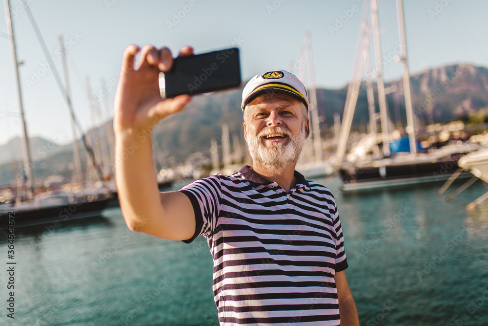 Mature man stands on pier dressed in a sailor's shirt and hat using smart phone to make photo.