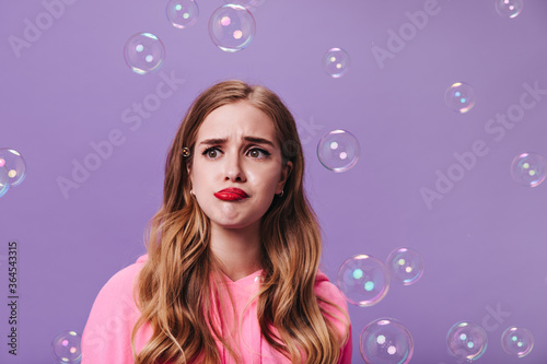 Charming girl in pink hoodie looking at bubbles on purple background. Blonde curly woman in street style outfit posing on isolated backdrop