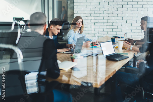 Team of multicultural male and female professionals dressed in formal wear discussing productive strategy together with proud ceo sitting at meeting table in stylish office interior behind glass wall photo