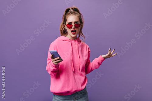 Girl in pink hoodie holding phone and looking into camera with misunderstanding. Pretty woman in street style sweatshirt looks frustrated on purple background