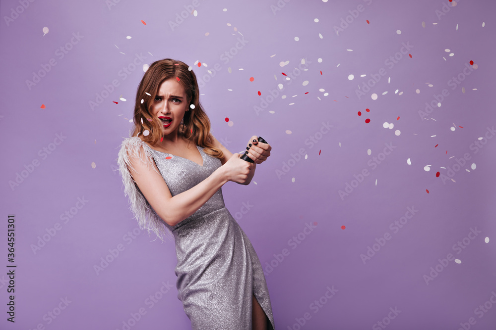 Blonde lady in silver dress throwing confetti. Portrait of curly attractive woman in festive outfit looking into camera and having fun on purple background