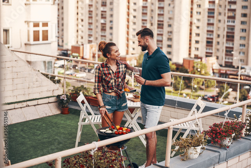 Charming young couple in casual clothing preparing barbecue and smiling while standing on the rooftop patio outdoors