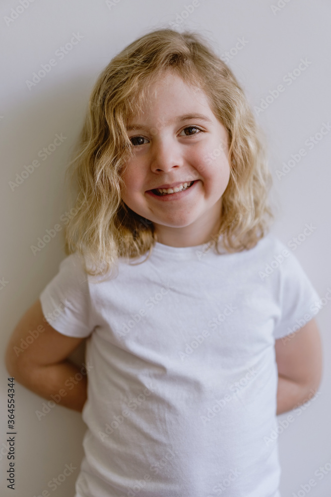 Positive child wearing casual clothes smiling and looking at camera while standing and leaning on white wall in modern studio Positive child wearing casual clothes smiling and looking at camera while standing and leaning on white wall in modern studio