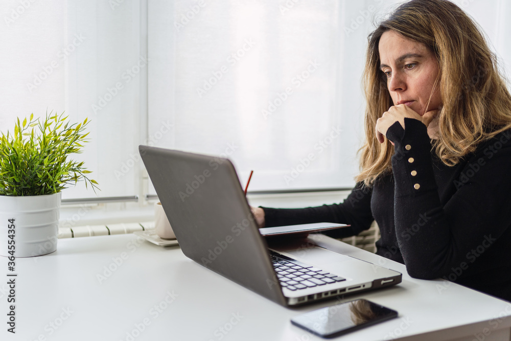 Side view of focused young female in casual outfit sitting at table with laptop and taking notes in planner while working online in light workplace at home Side view of focused young female in casual outfit sitting at table with laptop and taking notes in planner while working online in light workplace at home