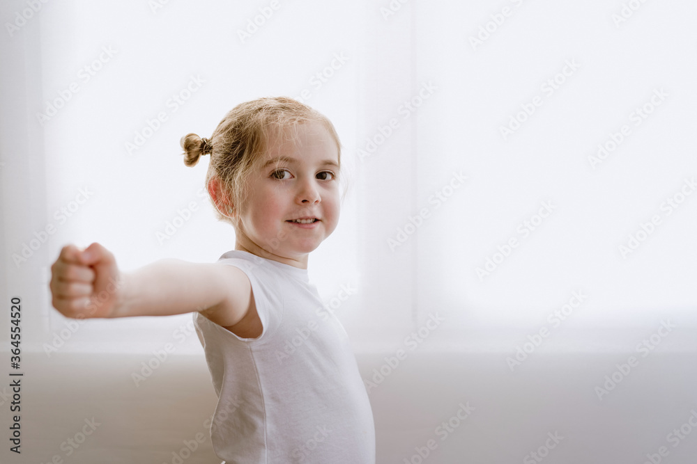 Side view of content little kid in casual outfit standing in bright apartment and stretching arms during warm up while looking at camera
