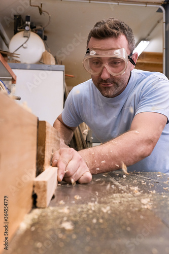 Low angle of focused carpenter in protective glasses milling timber while crafting detail using electric machine in light modern workshop photo