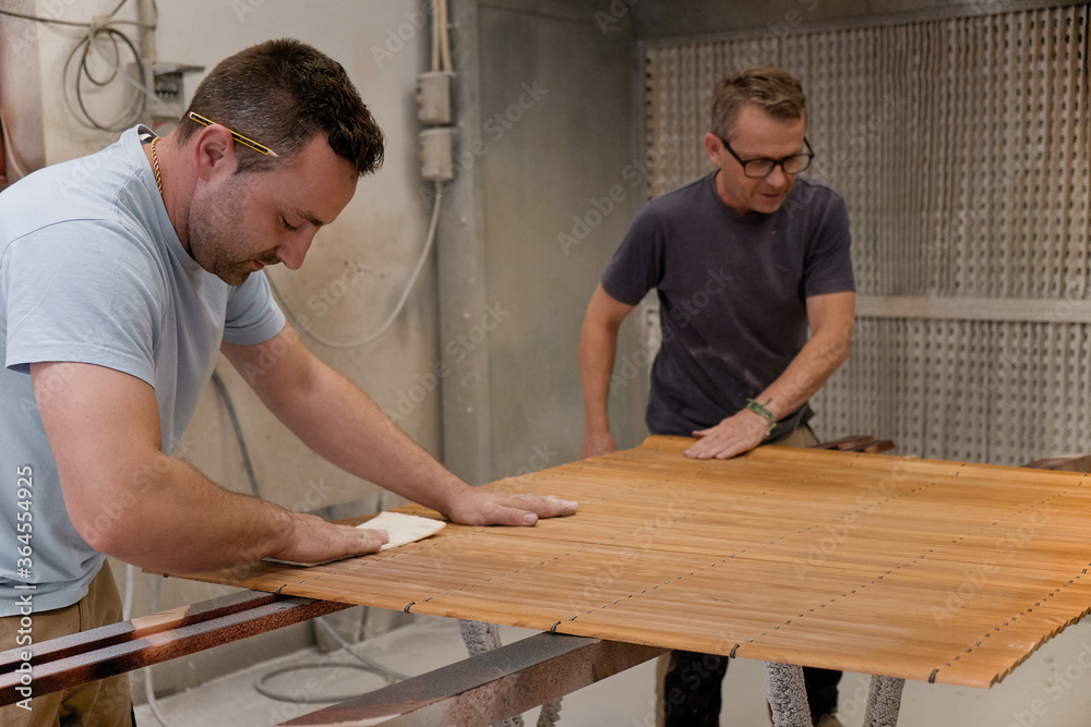 Adult focused man polishing wood slats painted with lacquer while working with colleague at rack in modern carpentry workroom Adult focused man polishing wood slats painted with lacquer while working with colleague at rack in modern carpentry workroom