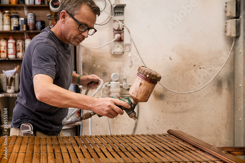 Craftsman applying varnish to wood jalousie using airbrush in carpentry workshop photo