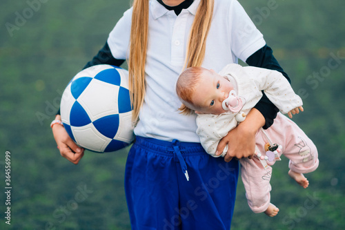 Unrecognizable young schoolgirl in white and blue uniform with soccer ball and doll on grassy field in sports club as concept for female choice and childhood photo