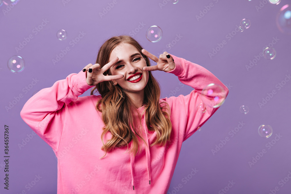 Happy lady in pink sweatshirt smiling and showing peace sign on purple background with bubbles. Joyful woman in hoodie laughing on isolated backdrop Happy lady in pink sweatshirt smiling and showing peace sign on purple background with bubbles. Joyful woman in hoodie laughing on isolated backdrop
