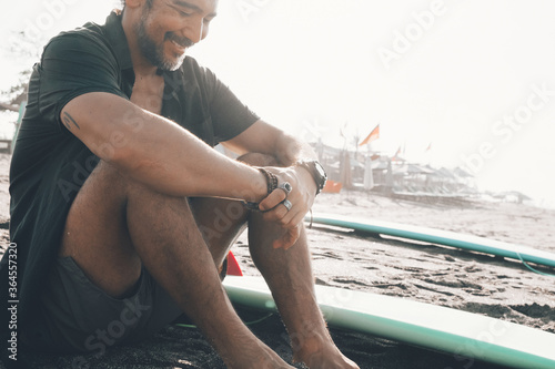 Positive young ethnic male in unbuttoned shirt and shorts enjoying summer day while resting on sandy beach near surfboard photo