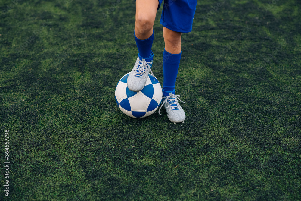High angle of faceless child in blue shorts and cleats with ball standing alone on green court while playing football at modern stadium High angle of faceless child in blue shorts and cleats with ball standing alone on green court while playing football at modern stadium