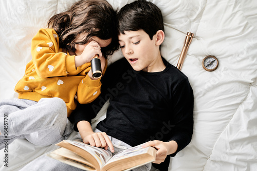 From above of brother reading book and sister playing with retro spyglass relaxing on comfortable bed and enjoying weekend photo