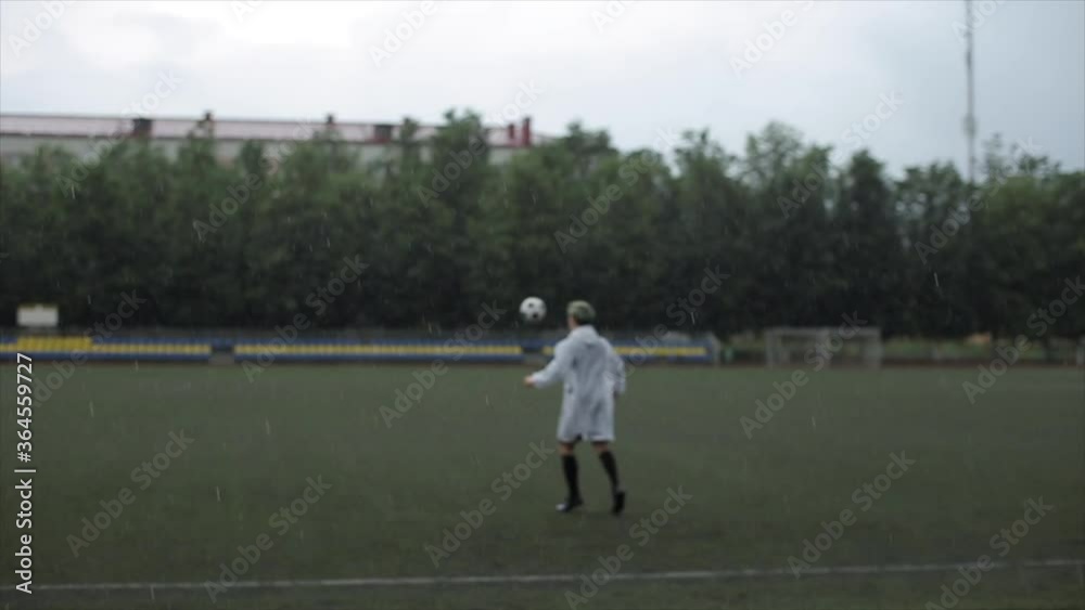A player of a soccer team in a raincoat is training on the field with the ball in the rain. The camera changes focus from a football player to raindrops A player of a soccer team in a raincoat is training on the field with the ball in the rain. The camera changes focus from a football player to raindrops