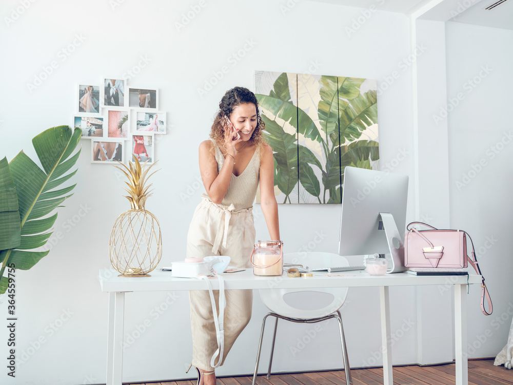 Pretty woman in elegant outfit smiling and having phone conversation while standing near desk in stylish office Pretty woman in elegant outfit smiling and having phone conversation while standing near desk in stylish office