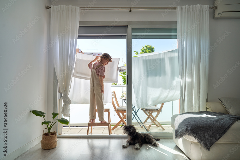 Back view of kid in casual clothes standing on chair on balcony and hanging wet linen on rope with dog lying down on the floor at home Back view of kid in casual clothes standing on chair on balcony and hanging wet linen on rope with dog lying down on the floor at home