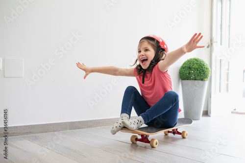 Little kid in red spotted helmet and casual clothes and dreaming about outdoors amusement while sitting alone on longboard in corridor of light modern apartment staying at home photo